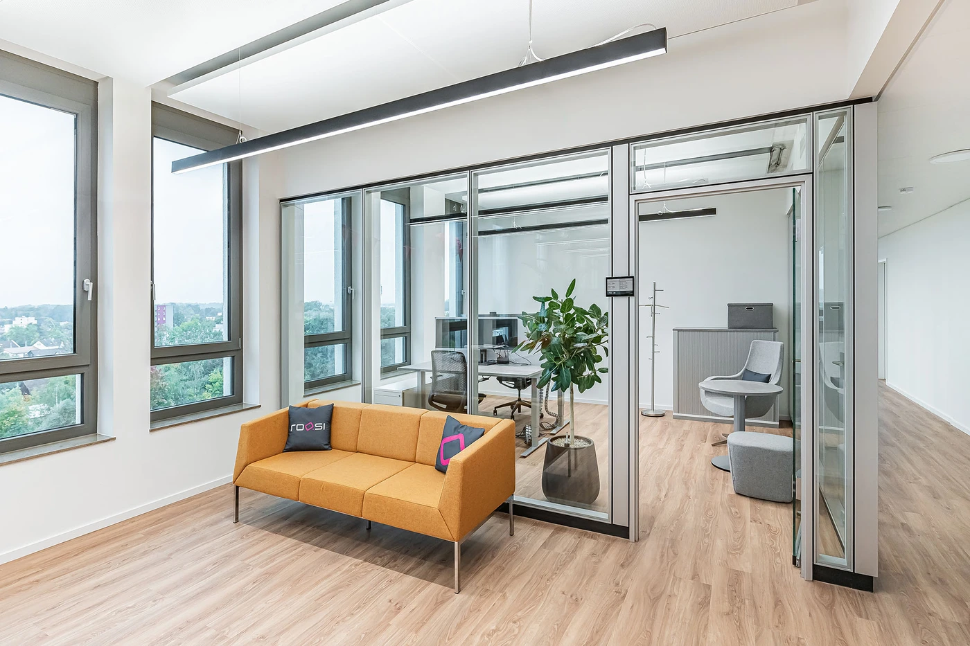 Loungebereich im roosi-Büro Rosenheim mit orangefarbenem Sofa und roosi-Kissen, große Fenster mit Blick ins Grüne